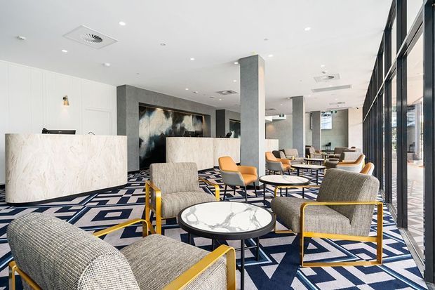 A bright, contemporary commercial lounge or hotel lobby with floor-to-ceiling glazing, white ceilings, grey stone-clad columns, sculptural reception desks, and multiple upholstered lounge chairs around round coffee tables. The space features a bold geometric navy, white, and grey carpet, brass-gold chair frames, and abstract wall artwork.