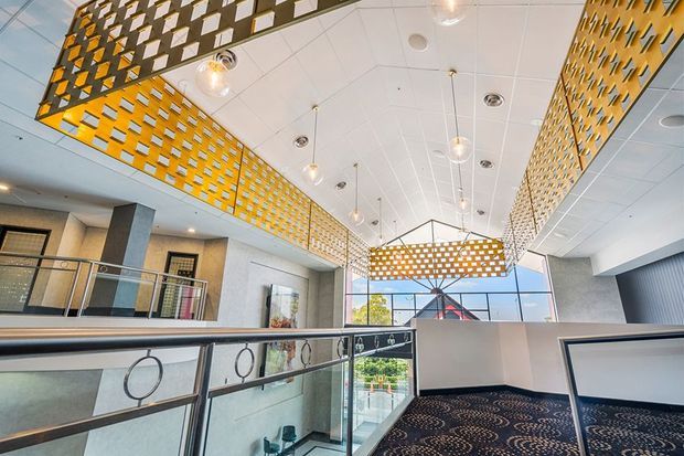 A bright contemporary commercial interior, likely a multi-level lobby or atrium, viewed from an upper-level walkway. The space features expansive glazing, patterned metal privacy screens forming suspended geometric ceiling elements, pendant globe lights, pale textured walls, dark carpet with circular motifs, and metal-and-glass balustrades with circular decorative details.