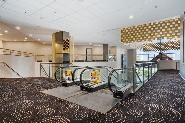 Bright contemporary commercial interior showing a pair of escalators descending between carpeted levels, with metal and glass balustrades, patterned carpet, open circulation areas, and a prominent perforated decorative ceiling feature above the escalator opening.