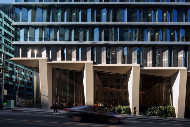 Street-level view of a contemporary multi-storey urban commercial building, featuring a strongly articulated glazed facade with repetitive floor-to-ceiling windows, dark vertical fins, projecting horizontal slab bands, and a monumental recessed ground-floor entrance framed by pale structural portals. Pedestrians and motion-blurred traffic appear in the foreground.