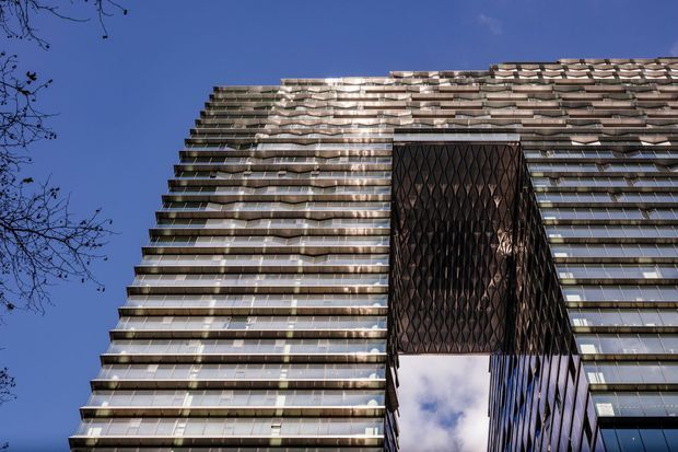 Upward-looking exterior view of a contemporary multi-storey building with extensive horizontal bands of glazing and projecting balconies or sunshade elements. A tall central opening is enclosed by a dark diamond-patterned structural screen, creating a dramatic portal through the building. The facade is predominantly metallic and glass, set against a clear blue sky.