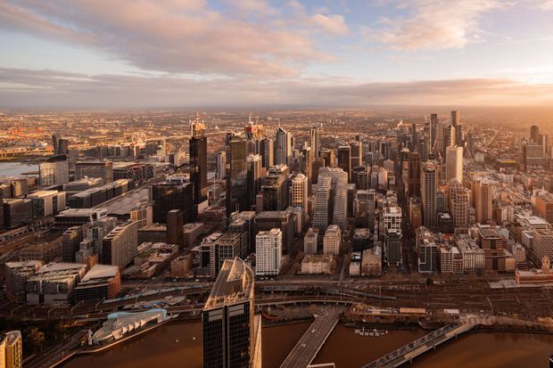 Aerial panoramic view of a dense modern city centre at sunset, with numerous high-rise office and residential towers, mid-rise commercial buildings, rail infrastructure, roads, and a waterfront or river edge in the foreground. The image is primarily an urban exterior cityscape rather than a single building or interior space.