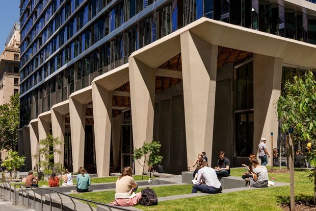 Exterior view of a contemporary multi-storey commercial or institutional building in an urban setting. The facade combines extensive dark reflective glazing with a rhythmic series of large, angled pale concrete portal-like frames forming a covered entry or arcade. Landscaped lawns, young trees, paving, steps, and people seated outside define the forecourt. The image emphasizes the building facade and public outdoor space.