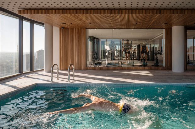 Indoor swimming pool with a swimmer in the foreground, surrounded by full-height glazing and warm vertical timber wall and ceiling slats. A gym is visible through the large internal window, while strong daylight and city views enter from the glazed facade. The space has a refined contemporary recreational amenity feel.