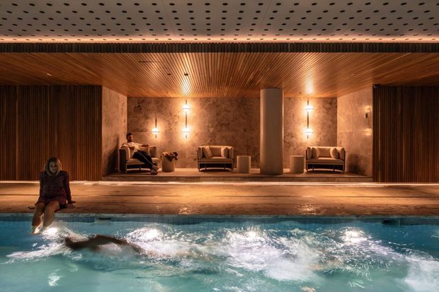 A sophisticated indoor pool and wellness lounge with warm timber-lined ceilings and walls, textured stone feature walls, integrated pendant and wall lighting, and built-in seating arranged along the pool edge. The foreground is dominated by a turquoise swimming pool with a person in the water; the lounge beyond has a calm, luxurious atmosphere.