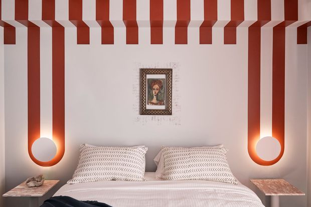 A stylized bedroom with a bed centered against a pale wall, flanked by dramatic red-and-white striped vertical wall treatment and matching sculptural pendant lights. Patterned pillows, warm bedside lighting, marble-look bedside tables, and a framed artwork create a bold, playful, graphic interior.