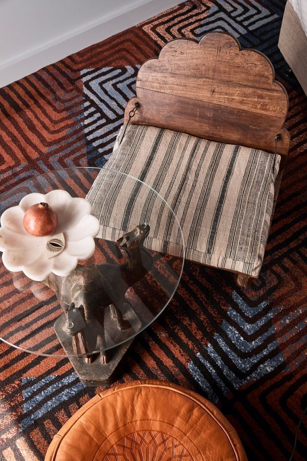 A close-up overhead view of a richly layered living room vignette: a vintage wooden lounge chair with a scalloped back and striped upholstered seat, a clear glass coffee table with a sculptural dark animal-shaped base, and a low rounded orange leather ottoman. A bold geometric patterned rug in rust, black, cream, and blue covers the floor. The composition has a warm, eclectic retro character.