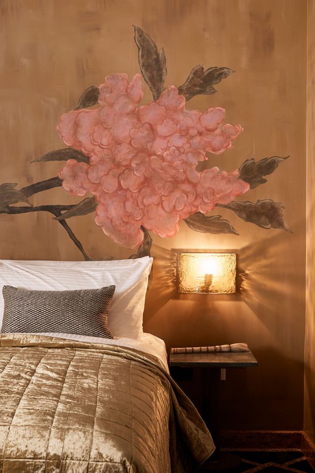 Close-up view of a warmly lit bedroom corner featuring a bed with layered white and taupe bedding, a dark textured accent pillow, and a small bedside table. The timber-toned wall is dominated by a large pink floral mural or artwork, with a glowing wall-mounted bedside lamp creating a cozy, luxurious atmosphere. A patterned dark rug is partially visible at the floor.