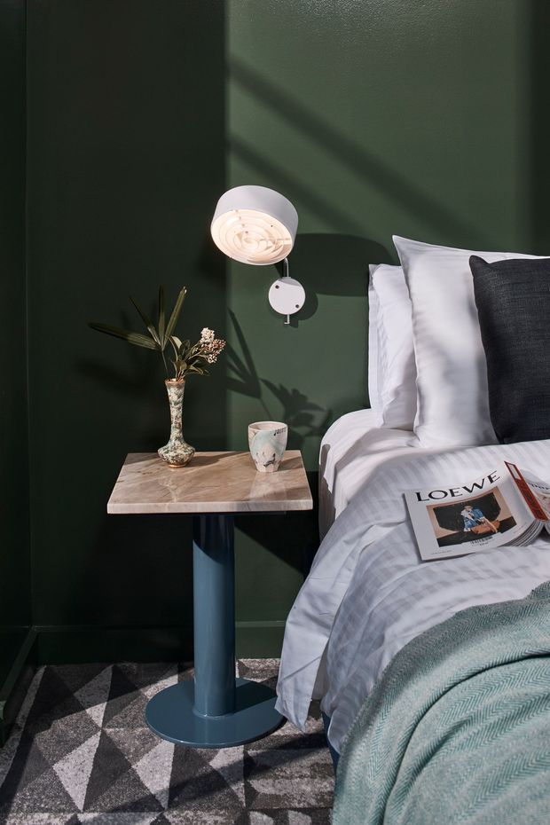 A moody, styled bedroom corner featuring a bed with white and pale grey bedding, a dark charcoal cushion, and a textured sage-green throw. A compact marble-look bedside table with a blue cylindrical pedestal supports a decorative vase and mug, while a wall-mounted white reading lamp illuminates the deep green wall. A geometric black, grey, and white rug covers the floor, and a magazine rests on the bed.