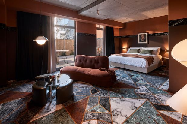 A moody, richly layered bedroom with a large upholstered bed, sculptural rust-coloured chaise sofa, round glass coffee table, dark curtains, bedside lighting, and a bold geometric patterned rug. The room combines terracotta walls, dark timber-toned surfaces, black marble-like accents, and an exposed concrete ceiling with visible structural beams. Natural light enters through a tall window and door opening, balancing the dramatic warm interior palette.