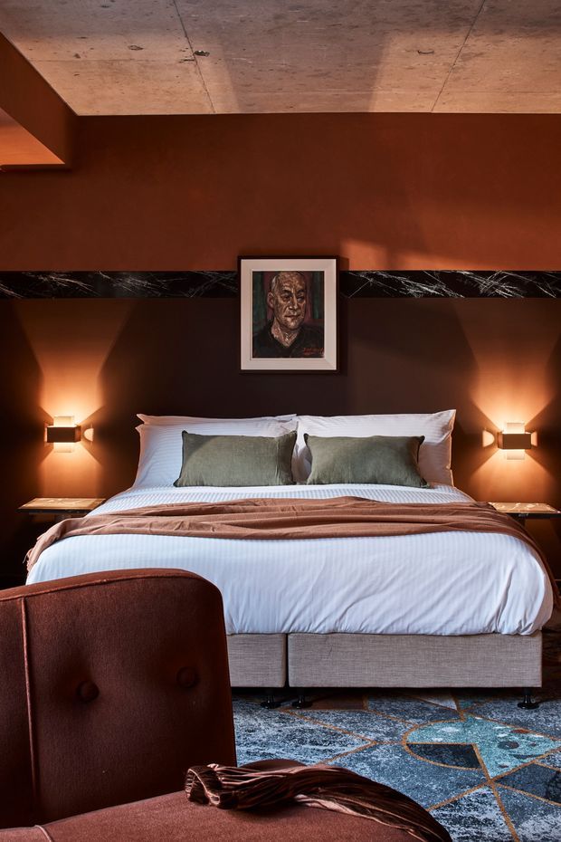 A warmly lit, richly layered bedroom featuring a large upholstered bed with white bedding, muted green cushions, and a rust-brown throw. Dark brown walls are accented by a horizontal black marble band with white veining, while wall-mounted sconces cast dramatic pools of light. A concrete-look ceiling, framed portrait, patterned blue-and-rust rug, and leather lounge chair complete the moody, refined interior.