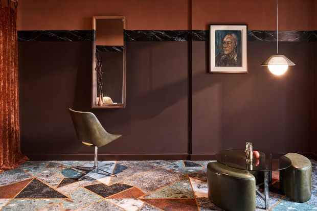 A richly styled compact sitting area with deep brown wall panels, a dramatic black marble horizontal trim, a sculptural lounge chair, full-height mirror, framed artwork, pendant lighting, and a geometric multicoloured patterned floor covering. A small dark round table with curved upholstered seating occupies the foreground. The interior has a moody, artful, high-contrast character.