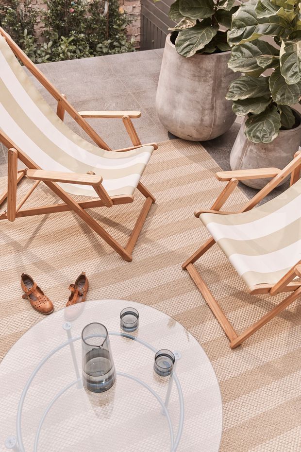 A styled outdoor terrace or courtyard seating area viewed from above, with several wooden-framed striped canvas sling chairs arranged around a round glass coffee table. Large potted plants, a textured beige outdoor rug, and sandals create a relaxed, natural entertaining setting. The palette is warm and muted, with timber, cream, beige, and leafy green tones.