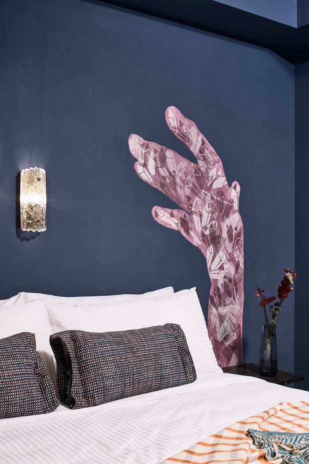 A stylised bedroom interior with a dark navy-blue wall, white bed linens, patterned dark throw pillows, and a large pink faceted botanical wall artwork behind the bed. A decorative wall sconce and a small bedside table with a vase add detail. The room has a dramatic, eclectic decorative character.