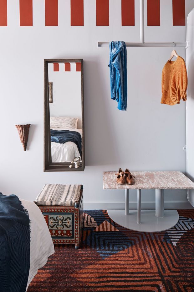 A compact, boldly styled bedroom with pale grey walls, a graphic red-and-white painted wall treatment near the ceiling, and a dark-framed full-length mirror reflecting the bed. A horizontal clothing rail holds blue and orange garments, while an ornate upholstered bench sits beside a slim stone-topped table. The room features a richly patterned geometric rug in rust, navy, blue, and cream tones, creating an eclectic and colourful atmosphere.