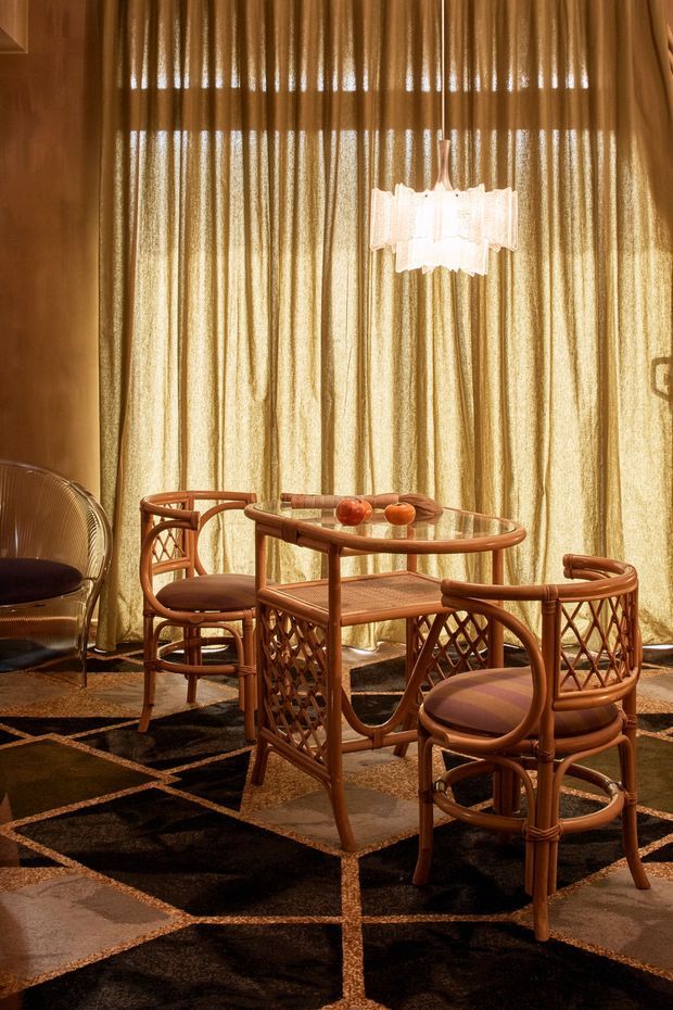 A warmly lit dining area with a small round glass-top dining table and four curved woven timber or rattan chairs. Floor-to-ceiling vertically textured golden curtains form the backdrop, with a sculptural pendant light above and a bold geometric dark patterned carpet underfoot. The room has an eclectic vintage character with strong natural-material detailing and rich amber-brown tones.