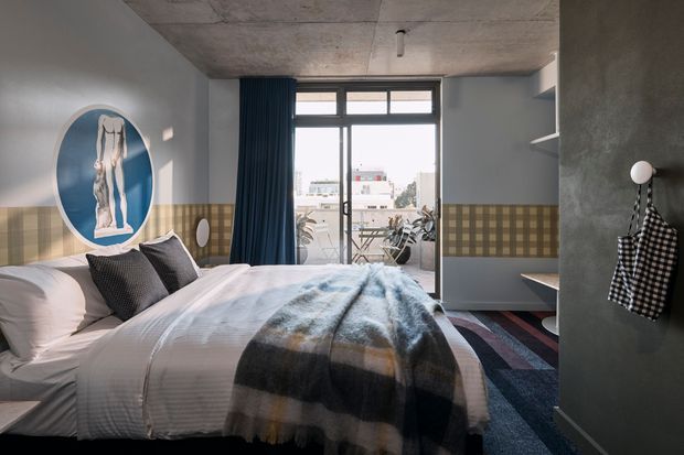 A contemporary bedroom with a large bed dressed in white bedding and a blue, grey, and mustard striped throw. The room features a grey concrete ceiling, muted grey walls, a patterned tile wainscot, dark blue curtains, built-in shelving, and a full-height glazed door opening to a balcony. A round artwork is mounted above the tiled wall, and a checkered garment hangs on a wall hook.