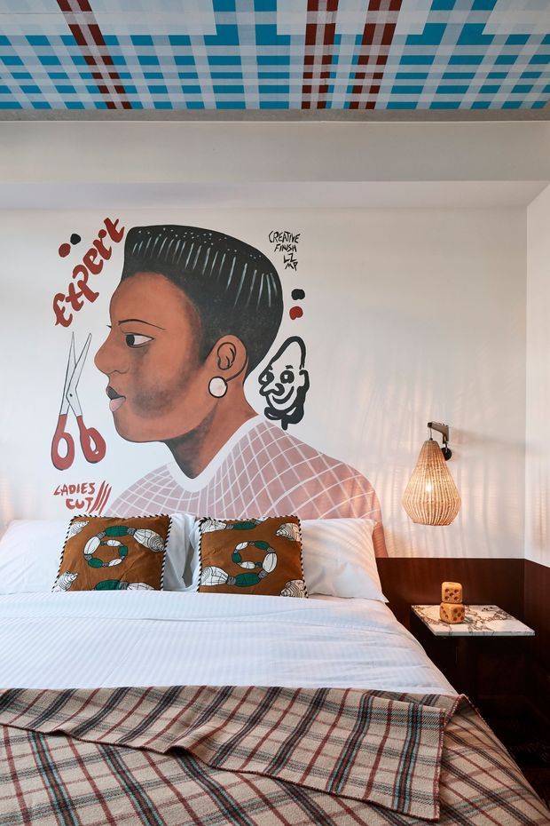 A vibrant, eclectic bedroom with a neatly made bed, patterned brown and teal cushions, and a plaid throw. The white wall behind the bed features a large hand-painted mural of a barbered profile with scissors and expressive lettering. A woven pendant lamp hangs beside the bed above a dark timber bedside surface, while the ceiling has a bold blue, white, and red geometric grid pattern.