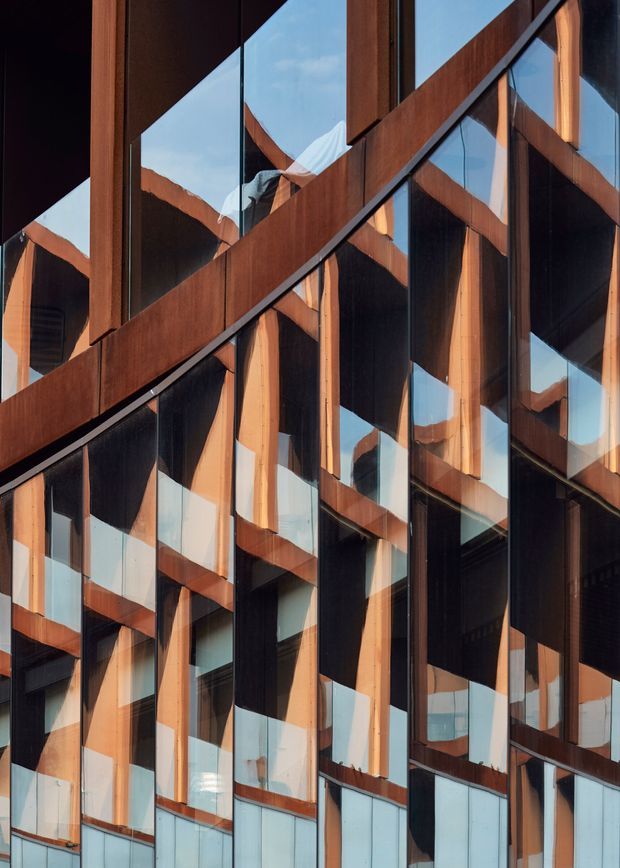 Close-up view of a contemporary building facade composed of extensive reflective glazing, dark vertical panels, and prominent warm-toned timber structural or framing elements. The irregular projecting and receding geometry creates a strongly angular, faceted pattern, with blue sky and surrounding forms reflected in the glass.