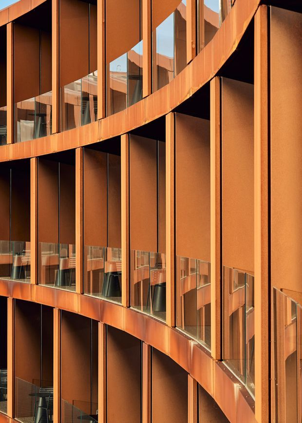 Close-up architectural view of a multi-storey building facade with curved bands of floor-to-ceiling glazing, vertical metal fins, and warm rust-coloured metal cladding. Reflections reveal repeated balconies or internal circulation spaces, creating a highly geometric, layered composition with strong vertical and horizontal lines.