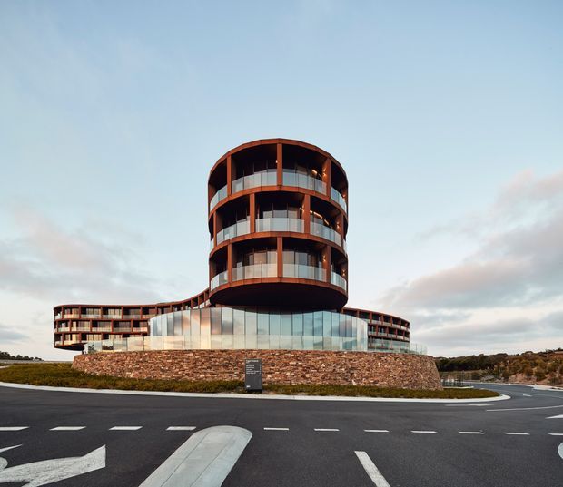 Exterior architectural photograph of a distinctive contemporary multi-storey building with a rounded central tower and curved wings. The structure features extensive floor-to-ceiling glazing, dark metal or timber-toned vertical framing, wraparound balconies with glass balustrades, and a broad stone-clad retaining wall at its base. It is viewed from a paved roadway under a bright sky.
