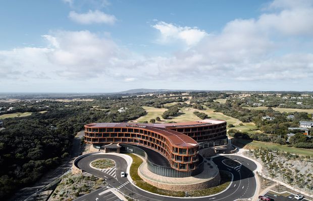 Aerial view of a large contemporary curved building set within an expansive landscaped and semi-rural coastal-looking site. The multi-storey structure has a rounded, ribbon-like form with extensive glazing, warm terracotta or copper-toned cladding, rooftop plantings, and a circular driveway with landscaped islands. Surrounding vegetation, open fields, roads, and distant development are visible under a bright partly cloudy sky.