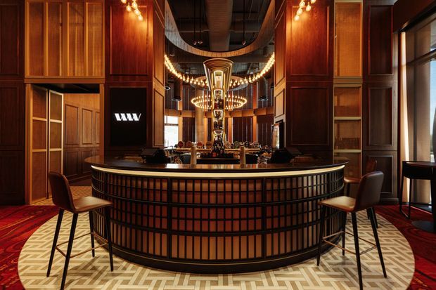 A dramatic upscale bar or reception counter dominates the interior, clad in dark timber panelling with a curved ribbed and gridded front. Two bar stools sit on a geometric patterned floor, while mirrored surfaces, warm pendant and globe lighting, dark ceilings, and rich red carpet create an opulent hospitality atmosphere.