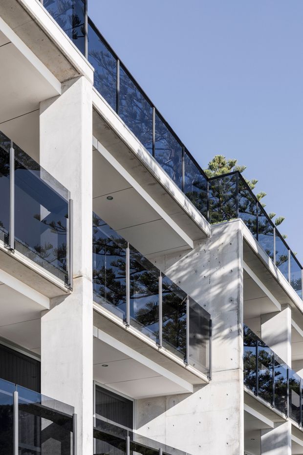 Close-up exterior view of a contemporary multi-level building with projecting concrete balconies, white concrete structural walls and columns, and transparent glass balustrades with dark metal frames. Strong geometric lines, recessed soffits, and reflected trees define the facade against a clear blue sky.