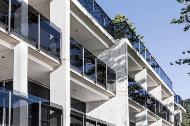 Exterior architectural photograph of a contemporary multi-storey residential building with strong geometric concrete forms, projecting balconies, extensive dark-framed glazing, and transparent glass balustrades. The facade is predominantly white and concrete, with repeated horizontal balcony lines and a clear blue sky visible around the structure.
