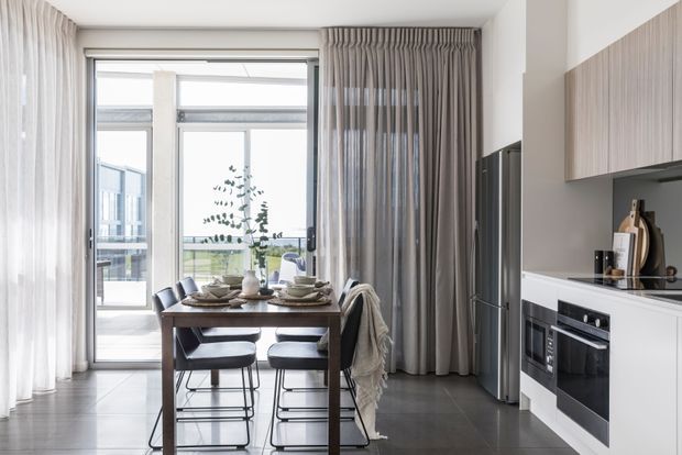 Bright contemporary open-plan dining and kitchen interior with a compact dark timber dining table, modern upholstered bar-style chairs, full-height sheer and grey curtains, dark tiled flooring, white cabinetry, a wood-look overhead cabinet, integrated black appliances, and floor-to-ceiling glazing opening toward a balcony or outdoor view.