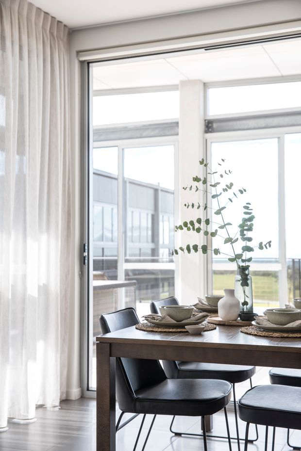Bright, refined dining area with a dark timber dining table and black upholstered chairs positioned beside expansive floor-to-ceiling glazing. Sheer curtains, pale flooring, neutral ceramic tableware, and a tall eucalyptus arrangement create an airy, contemporary setting. An exterior balcony or terrace is visible beyond the windows.