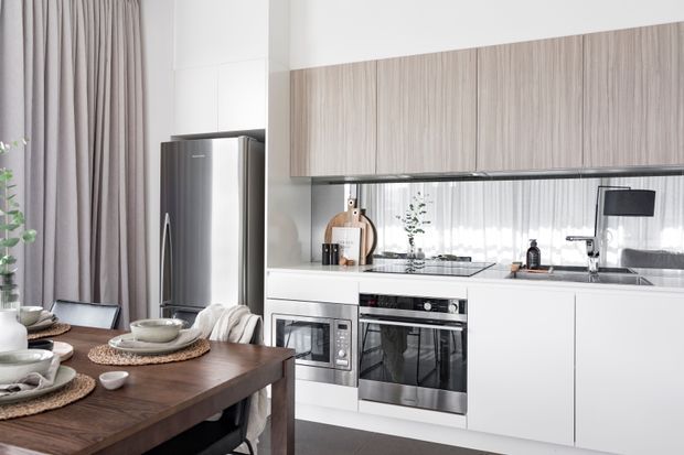 Bright contemporary open-plan kitchen and dining area with white flat-panel cabinetry, a light wood-grain overhead cabinet bank, stainless-steel appliances, mirrored splashback, timber dining table, and full-height taupe curtains. The kitchen includes an integrated oven and microwave, induction cooktop, sink, and refrigerator, with the dining setting in the foreground.