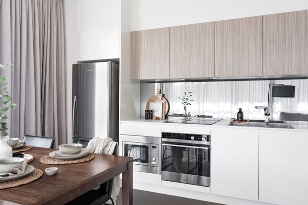 Bright contemporary open-plan kitchen and dining area with white handleless cabinetry, a long horizontal timber-look overhead cabinet, mirrored splashback, integrated stainless-steel appliances, and a dark timber dining table. Full-height taupe curtains and large glazing create a soft, airy atmosphere.
