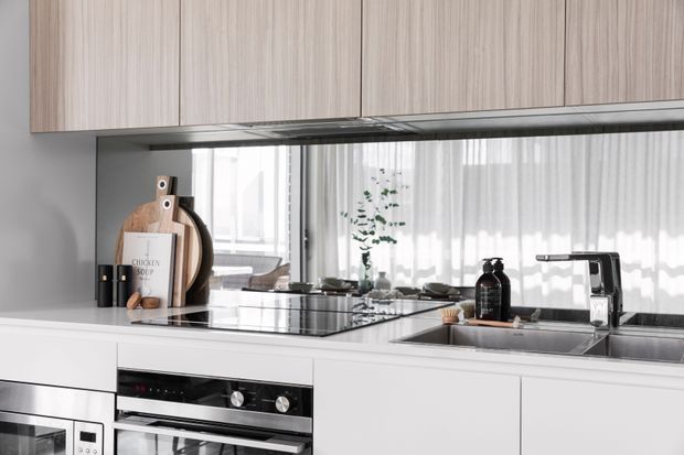 Contemporary kitchen with pale wood overhead cabinetry, white lower cabinetry, a dark reflective benchtop, integrated cooking appliances, and a stainless-steel sink beneath a mirrored splashback. The mirror reflects bright windows and sheer curtains, creating an airy, light-filled appearance.