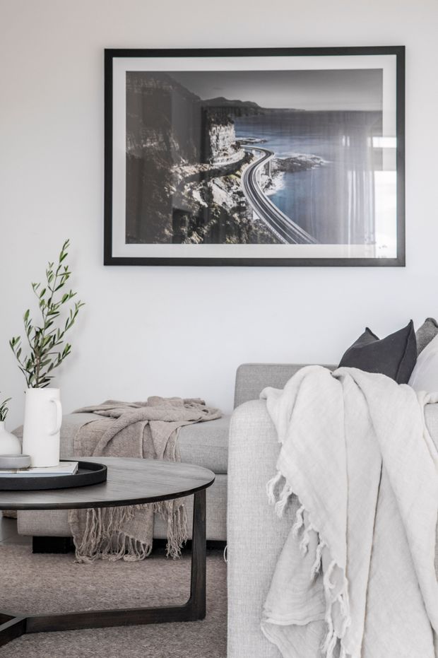 A softly styled living room with a neutral grey and white palette. The scene features a light grey upholstered sofa and armchair draped with textured throws, a dark round coffee table, a simple plant in a white vessel, and a large black-framed monochrome coastal road photograph on the wall. The overall look is calm, minimal, and contemporary.