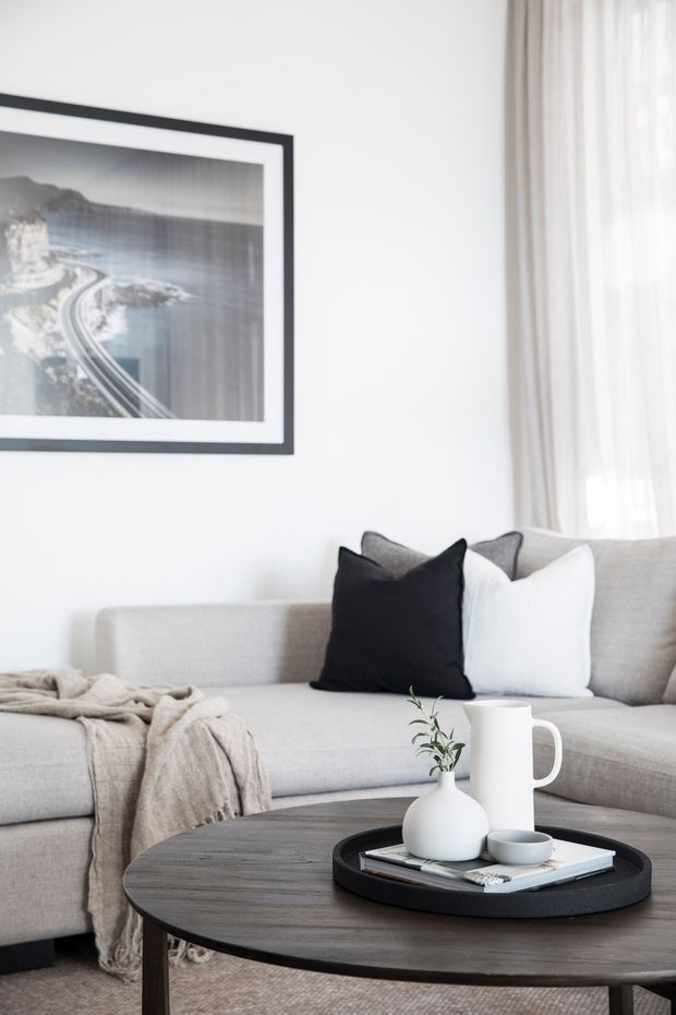 Bright, softly styled living room featuring a light grey sectional sofa with black, white and grey cushions, a neutral throw, a dark round coffee table with a tray and ceramic vessels, sheer floor-length curtains, and framed monochrome wall art. The overall composition is calm and contemporary with a restrained neutral palette.