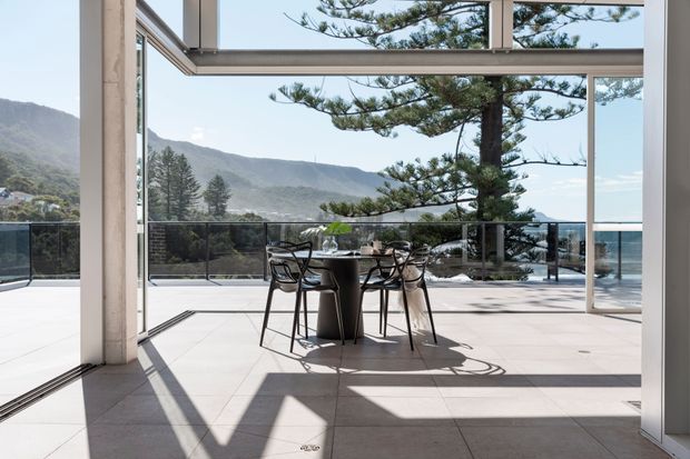 A bright, modern indoor-outdoor dining area with a small round table and chairs set on large pale tiled flooring. Full-height sliding glass doors open onto a spacious terrace with glass balustrades, framed by structural columns and expansive glazing. A mountain, coastal, or waterside landscape is visible beyond, with a large sculptural evergreen tree nearby.