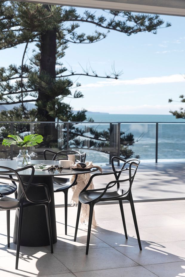 A bright, contemporary dining area opening onto a glass-balustraded terrace with expansive ocean views. A round black dining table is surrounded by sculptural black chairs, with pale large-format tiled flooring, extensive glazing, and a coastal landscape beyond.