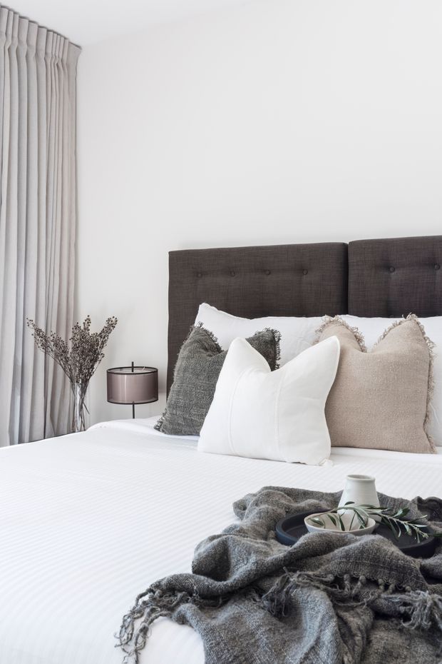 A softly styled bedroom featuring a large bed with a dark charcoal upholstered button-tufted headboard, layered white, grey, and beige pillows, and crisp white bedding. Floor-to-ceiling light grey curtains frame the left side, with a small bedside lamp and dried branches adding subtle decorative detail. A textured charcoal throw and tray with ceramics rest at the foot of the bed. The overall aesthetic is calm, neutral, and refined.