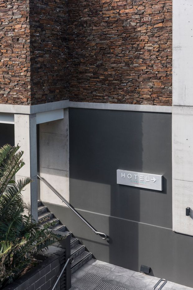 A contemporary hotel entrance stairwell with a dark rendered wall, exposed red-brown brick feature wall, raw concrete structural elements, and metal handrails. A wall-mounted sign identifies the space as a hotel. Tropical planting borders the stairs.