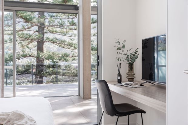 Bright, minimalist bedroom workspace with floor-to-ceiling glazing opening onto a balcony or terrace. A slim built-in desk with a large television, decorative vase, books, and a black chair sits beside the bed. Pale walls and large light stone or concrete floor tiles create a clean, contemporary interior, with trees visible outside.