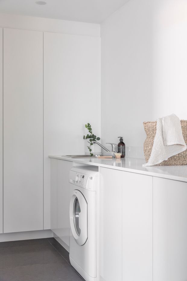 Bright minimalist laundry room with full-height white cabinetry, integrated front-loading washing machine, white countertop with inset sink and chrome tapware, and grey floor tiles. A woven basket and folded white towel add subtle texture.