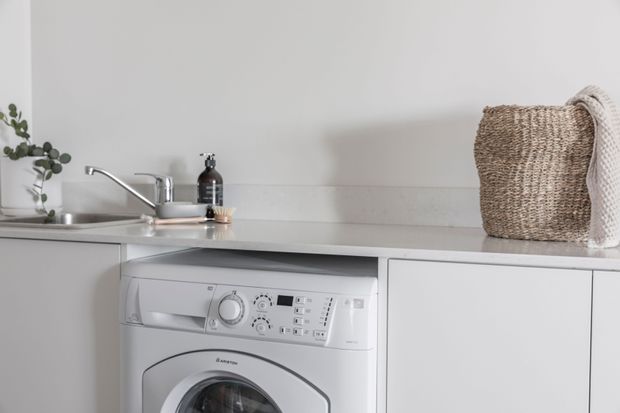 A bright, minimalist laundry area with a front-loading washing machine integrated beneath a pale benchtop. White flat-panel cabinetry, a stainless steel sink and tap, a light grey tiled or painted splashback, and a woven basket create a clean, neutral utility-space setting.