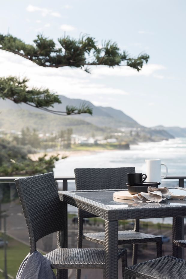 A furnished outdoor terrace or balcony overlooking a coastal bay, with dark woven outdoor dining chairs and table, place settings, and a glass balustrade. The background shows ocean, shoreline, and mountainous terrain, while sculptural evergreen branches frame the upper portion of the view.