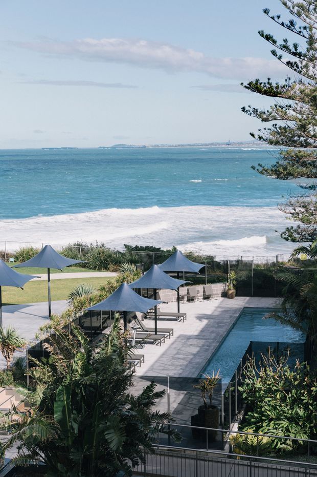 A coastal outdoor resort-like space overlooking the ocean, with a rectangular swimming pool, paved pool deck, dark shade umbrellas, lounge seating, landscaped tropical planting, and a sandy beach with waves beyond. A large coastal tree frames the right side of the view.