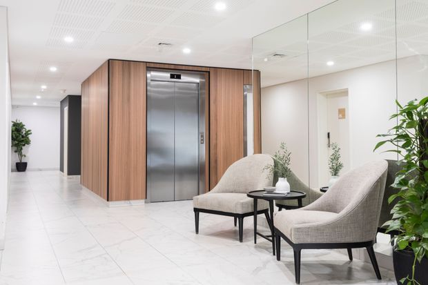 Bright contemporary commercial lobby or reception area with a stainless-steel elevator set into warm timber wall panelling. The space features polished white tiled flooring, a white perforated acoustic ceiling with recessed lighting, a large mirrored wall, and two upholstered lounge chairs with a small round side table and plants.