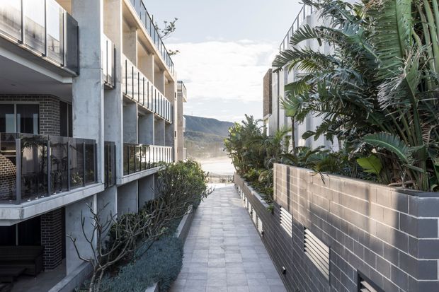 A landscaped paved walkway runs between contemporary multi-storey residential buildings, with balconies, dark brickwork, concrete structural elements and metal railings. Dense tropical planting lines the right side, while the path opens toward a distant mountainous coastal landscape.