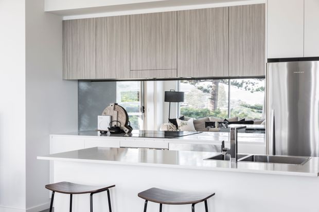 Bright contemporary kitchen with a large white island and integrated breakfast bar, dark wood stools, pale wood-grain overhead cabinetry, stainless-steel refrigerator, and a reflective splashback or mirrored wall opening toward a living area. Large windows provide abundant natural light.