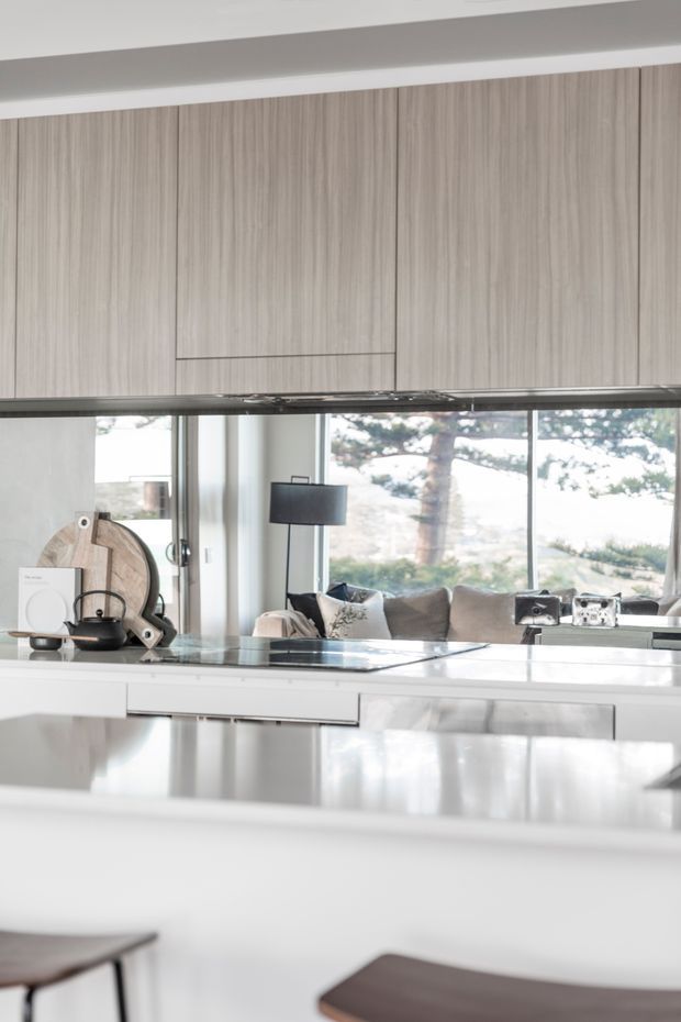 Bright contemporary kitchen with extensive pale timber overhead cabinetry, glossy white and stainless-steel lower cabinetry, and a reflective island or benchtop. Large glazing opens visually to a light-filled living area with a sofa and dark floor lamp, while a few kitchen appliances and accessories sit on the counter. The space has a clean, refined, neutral palette and strong indoor-outdoor connection.