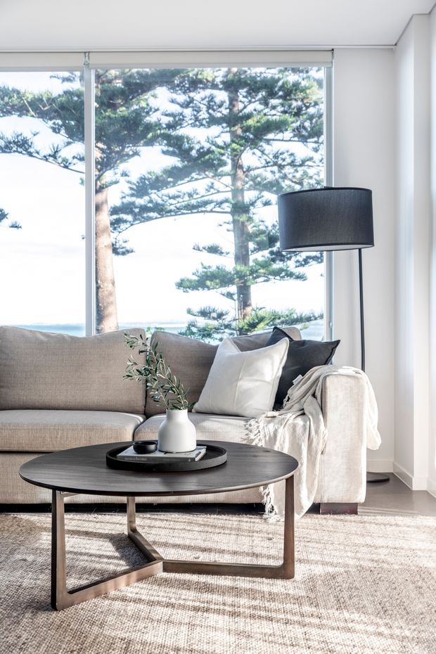 Bright, refined living room with a neutral grey-beige sectional sofa, layered cushions and throw, a round dark timber coffee table, black floor lamp, and simple ceramic vase. Full-height glazing provides views of mature coastal trees and water beyond. The space has a clean, contemporary feel with abundant natural light and textured soft furnishings.
